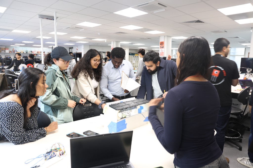 A group of students gathered around a hardware prototype on a lab bench.