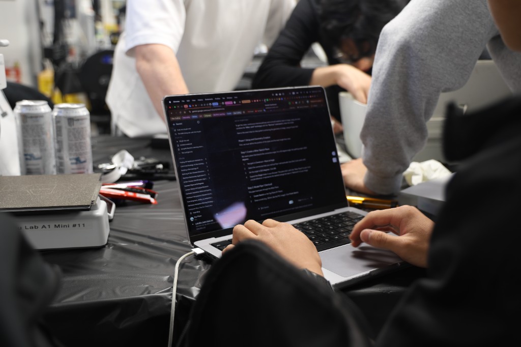 Hands on a laptop in a KCL hardware lab, with a 3D printer beside the keyboard.
